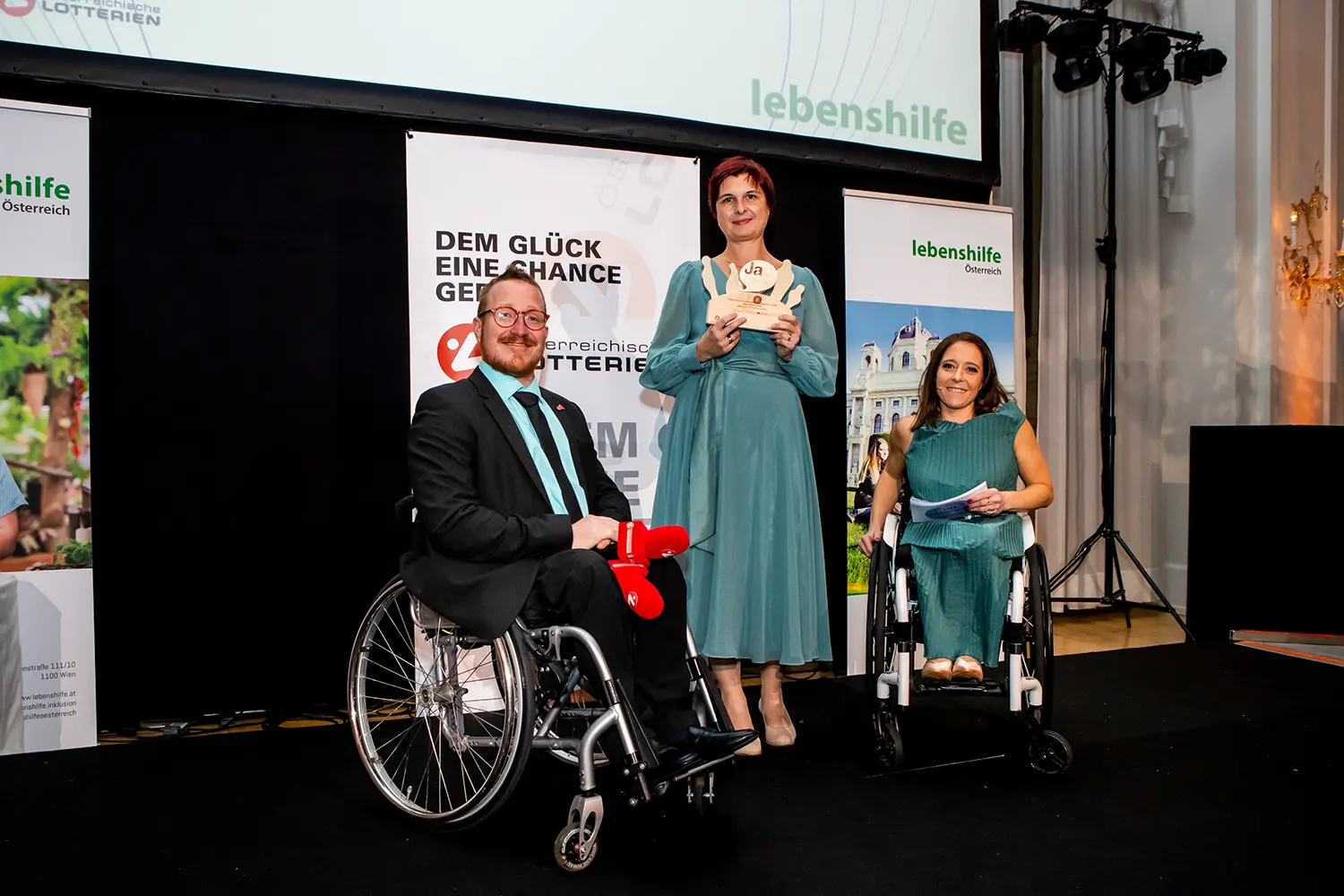 3 Menschen auf einer Bühne, Barbara Füreder-Kitzmüller in der Mitte mit dem Preis